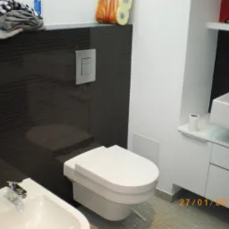 Nowoczesna łazienka z czarnymi, błyszczącymi płytkami na ścianie, wiszącą toaletą, bidetem z chromowaną baterią i białą umywalką nablatową na szafce.